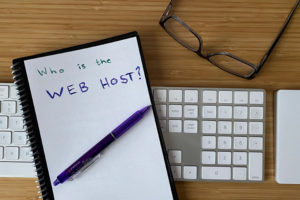A notebook with "who is the web host?" written on a page, resting on a computer keyboard